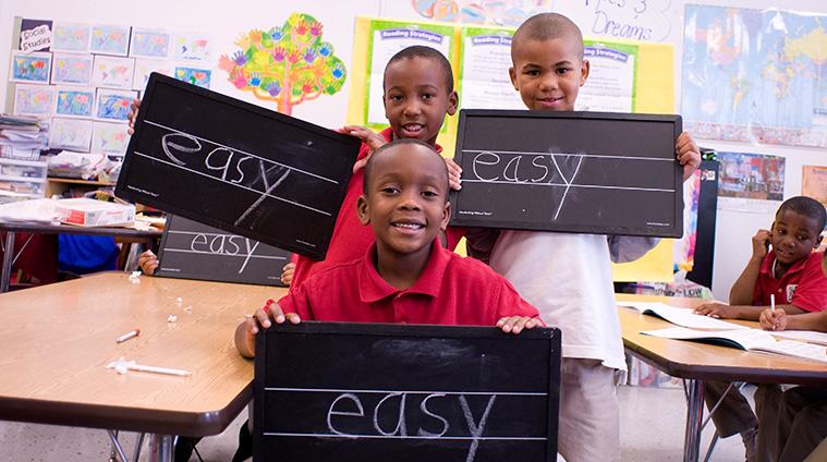 Three young students smiling and proudly holding blackboard signs that say 'EASY' in a classroom, representing the simplicity of the Learning Without Tears curriculum.
